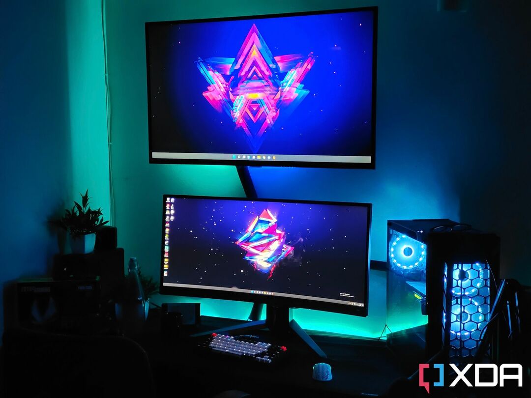 Vaizdas, kuriame rodoma žaidimų sąranka su televizoriumi, monitoriumi ir žaidimų kompiuterio dėklu ant stalo su RGB lemputėmis.