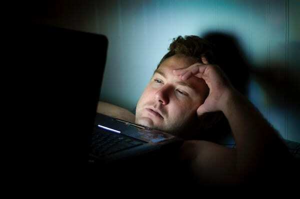 ノートパソコンの画面からの青い光で顔が照らされているベッドの男の写真